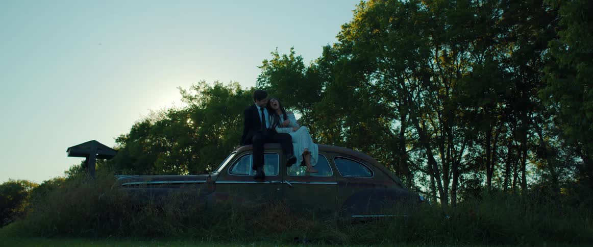 Newlyweds sit on the roof of a vintage car laughing and talking