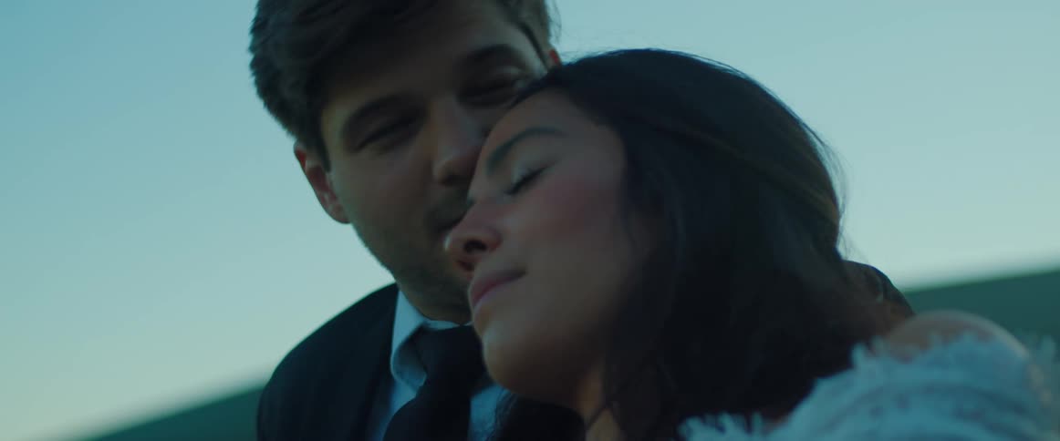 A man with a shirt and tie kissing a woman on her forehead with the sky and clouds in the background
