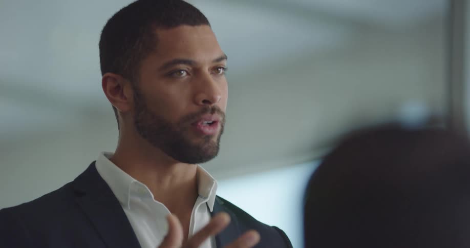 A black man stands in a room talking to other people