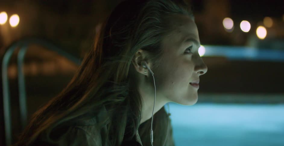 Woman wearing earphones sitting by a pool at night