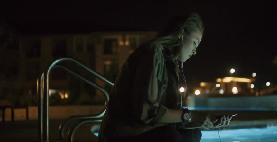 A woman sitting by a lit up pool at night plugs earbuds into her phone