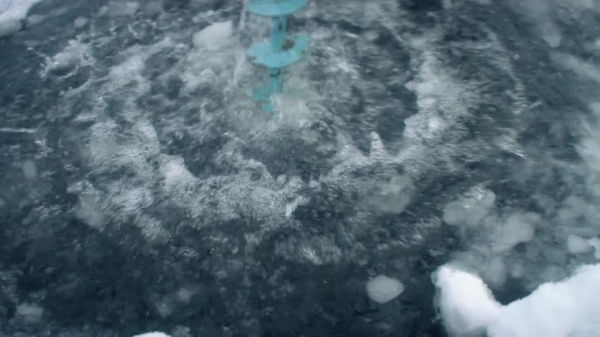 A person with a drill drilling a hole in the ice with snow and water