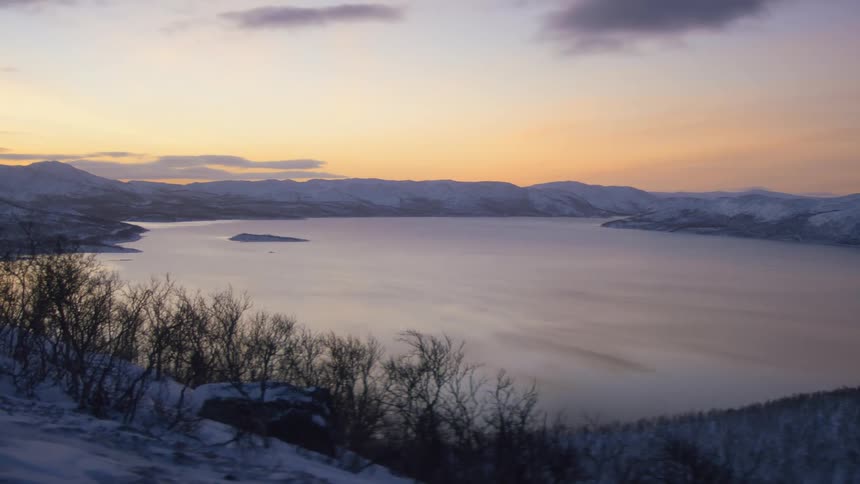 Sun rising in the distance behind a mountain range with a lake in front of it and snow covering the ground