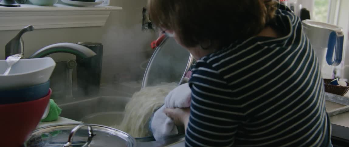 A woman cooking at home carries a pot of pasta to the sink and drains it