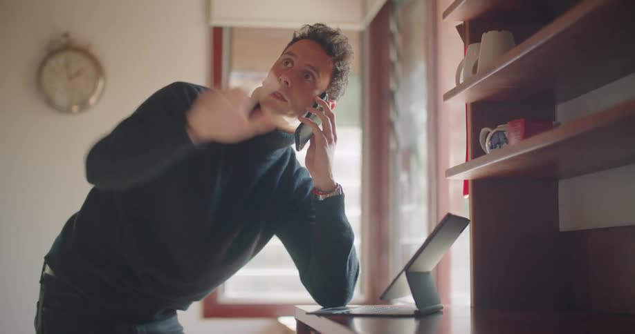 A man in a room with a smart phone using a laptop with a wall in the background