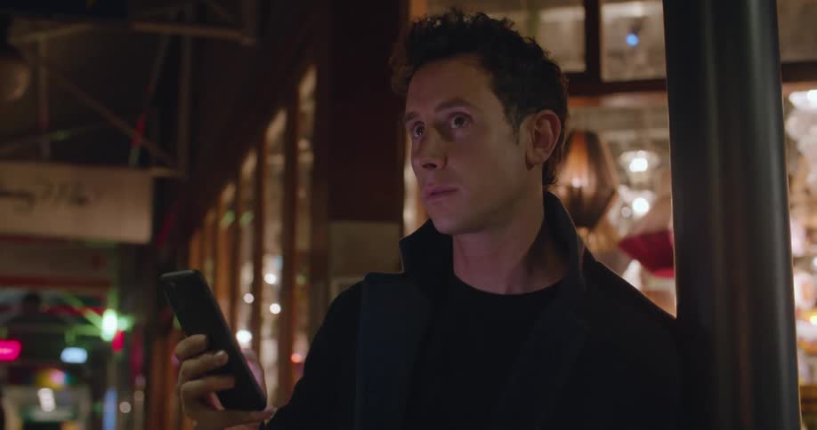 A man standing outside leaning against a pole scrolling on a smart phone and looking around