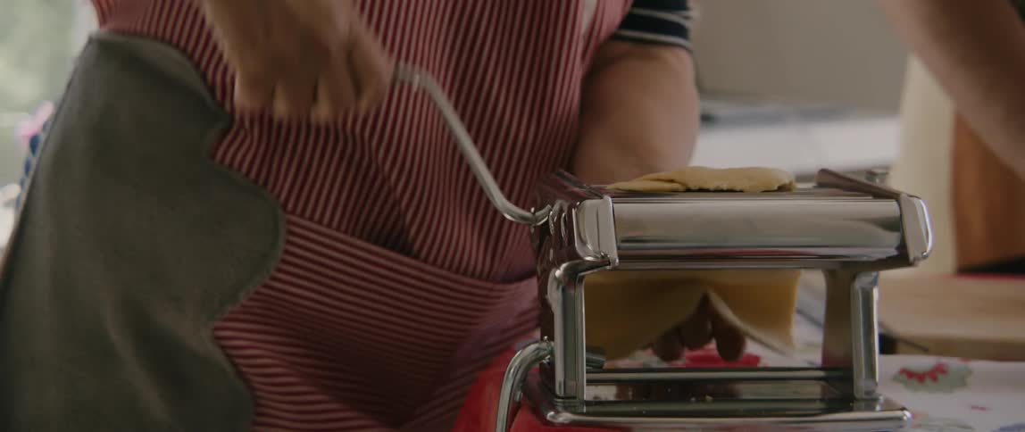 A person in a striped apron feeding dough through a pasta roller