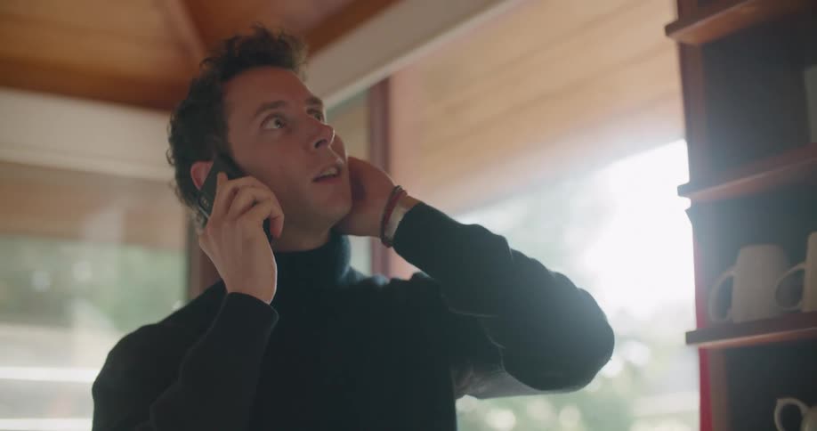 Man wearing turtleneck stands near shelf full of ceramic cups looking around while talking on mobile phone