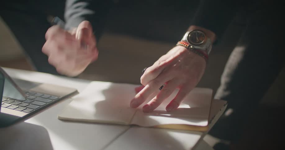 A person with a wristwatch using a pen to draw on a book on a table next to a laptop