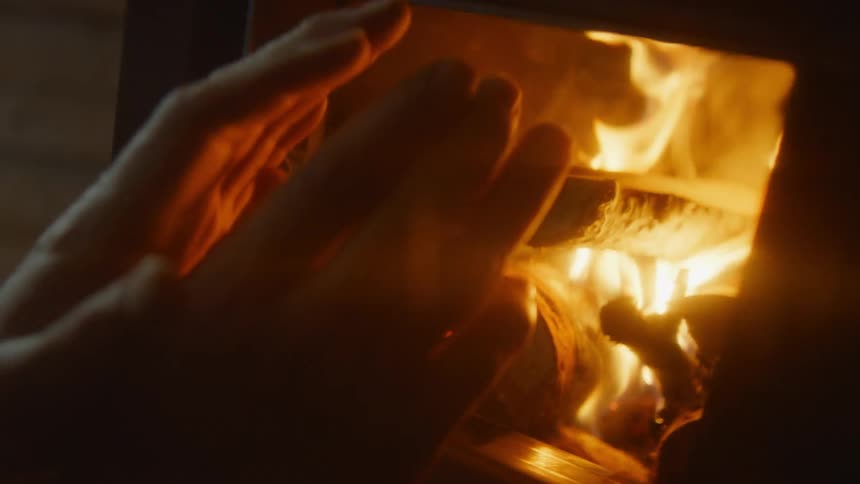 A man adds wood to a wood burning stove and uses it to warm his hands