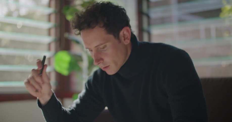 A man reads the screen of his laptop for a moment before writing in a notebook while sitting on a sofa indoors during the day