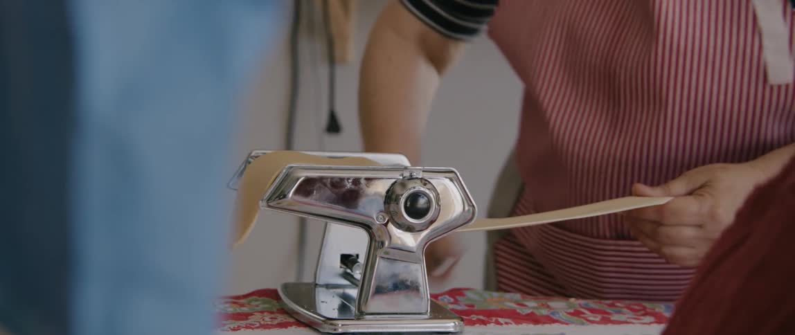 A woman uses a metal pasta maker to produce a flat piece of raw pasta to cut