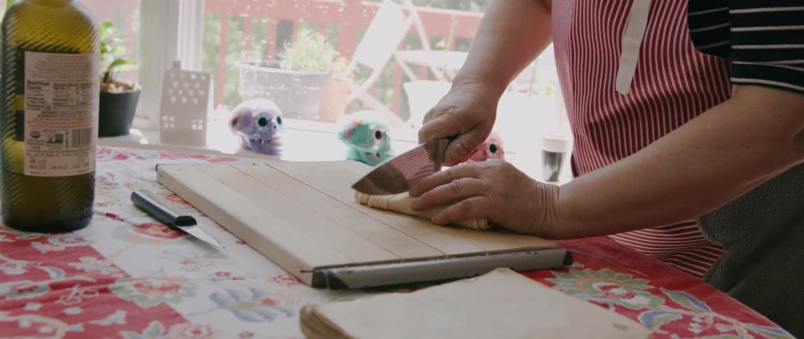 A woman slices a pastry with a large knife on a cutting board on top of a table indoors at home during the day