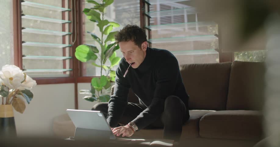 A man in a room using a laptop with a wall and window in the background