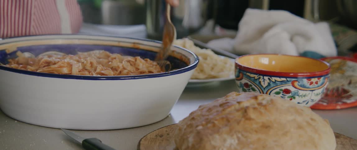 A woman uses two forks to mix pasta in a large bowl on a countertop next to other food items