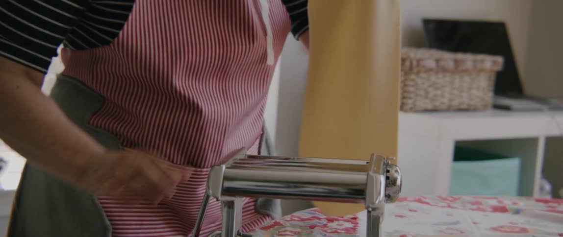 Woman runs dough through hand cranked pasta machine placing flattened sheet onto cloth lined table