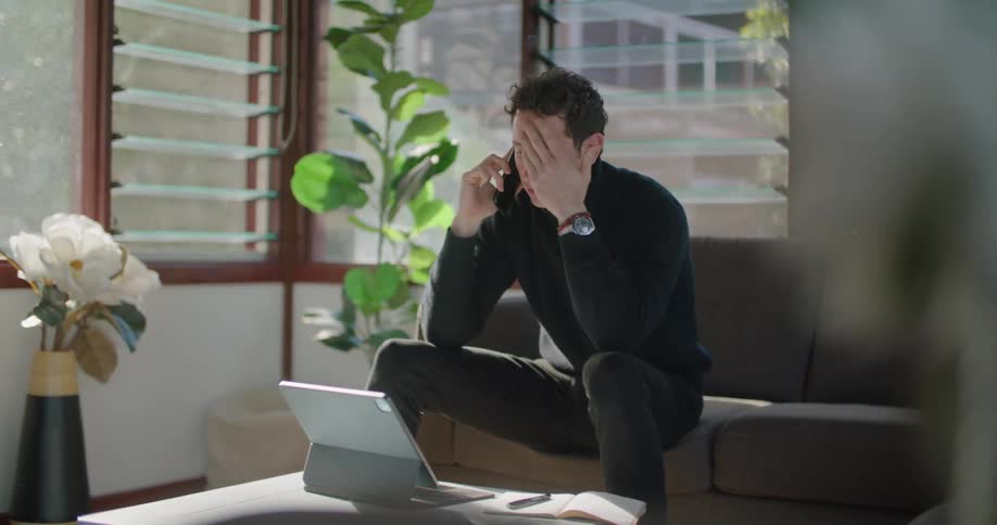 A man in a room sitting on a couch talking on a smartphone with a wall and window in the background