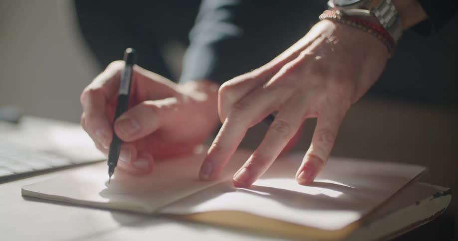 A person with a wristwatch writing on a book with a pen on a table