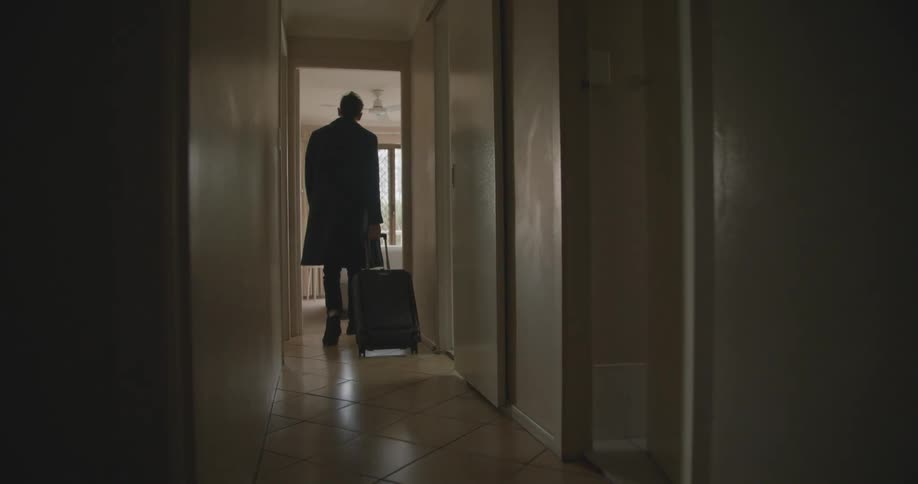 Man wearing heavy coat walks down narrow hallway pulling suitcase