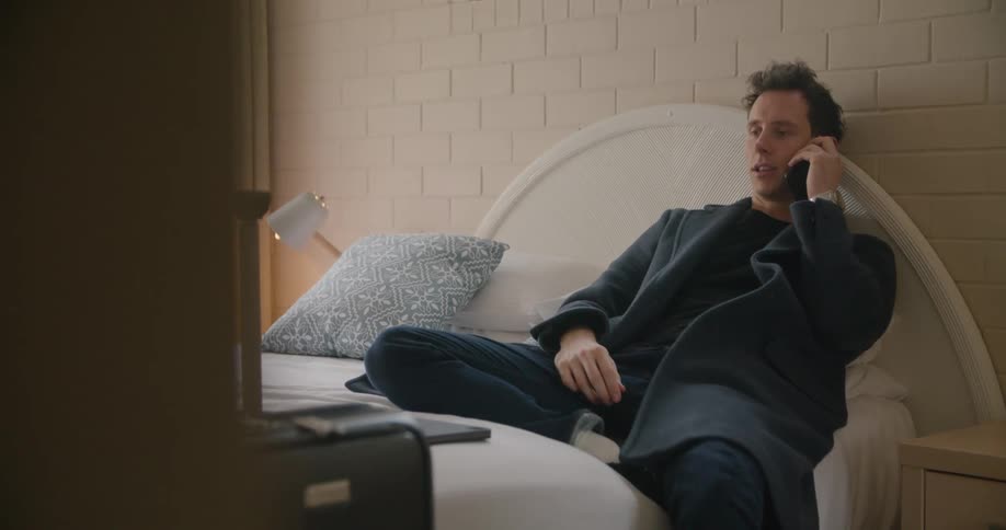 A man typing on a laptop in a hotel room leans back as he answers a phone call