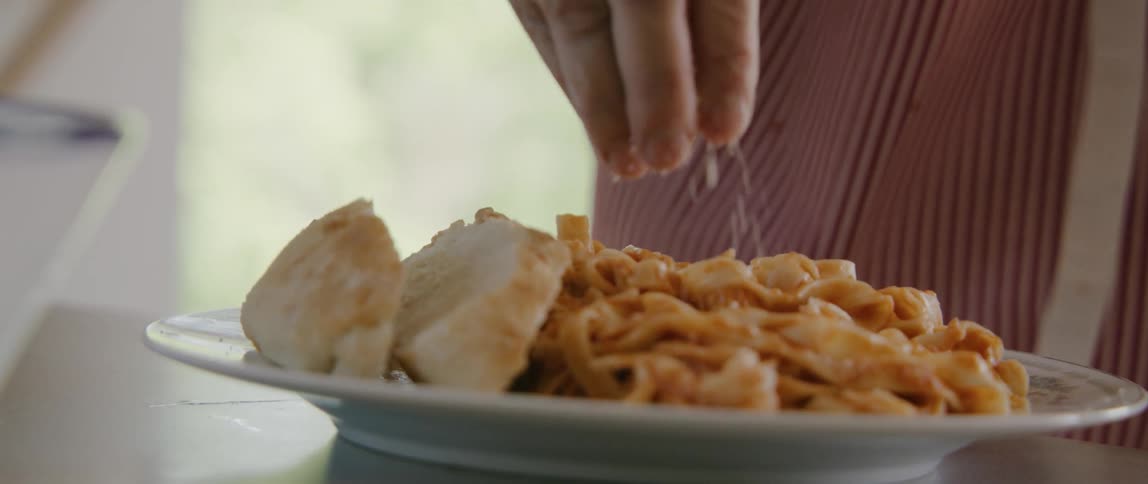 A person sprinkling parmesan on a plate of cooked pasta with bread