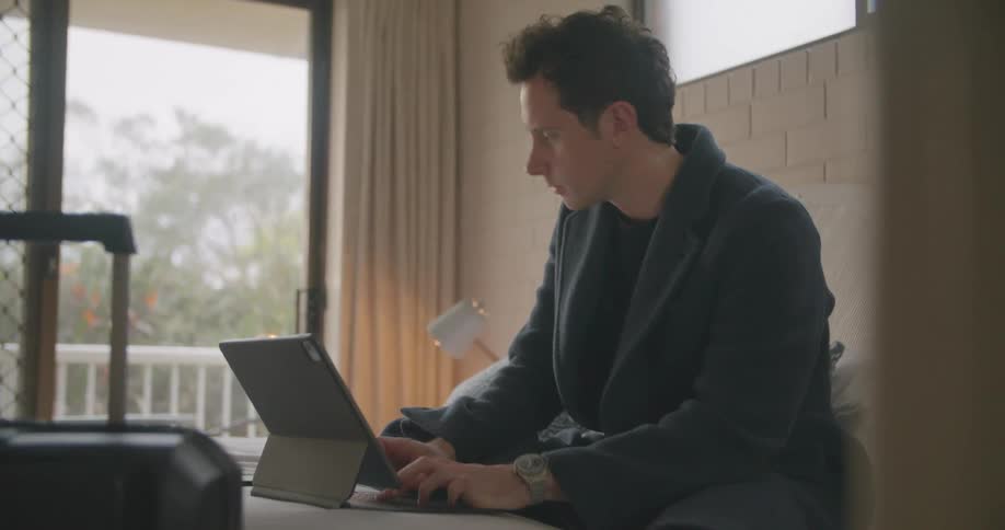 A man types on a laptop while sitting on a bed in a hotel during the day