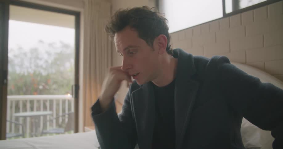 A man sits on a bed at home and video chats on a laptop