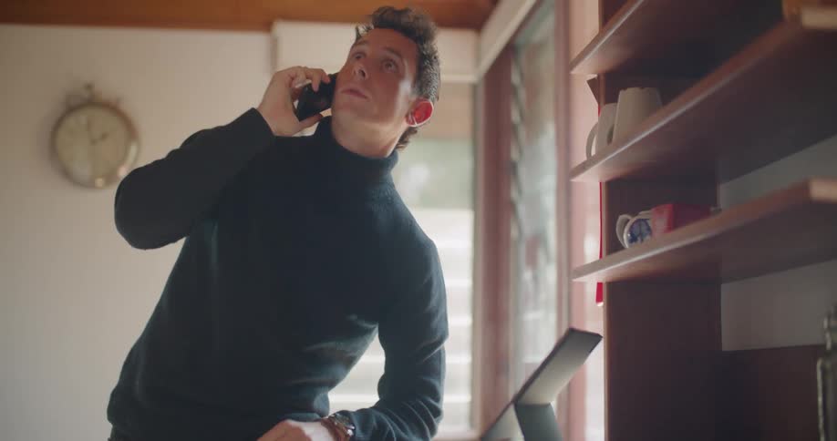 A man in a turtleneck sweater talking on a phone while looking at a laptop at home