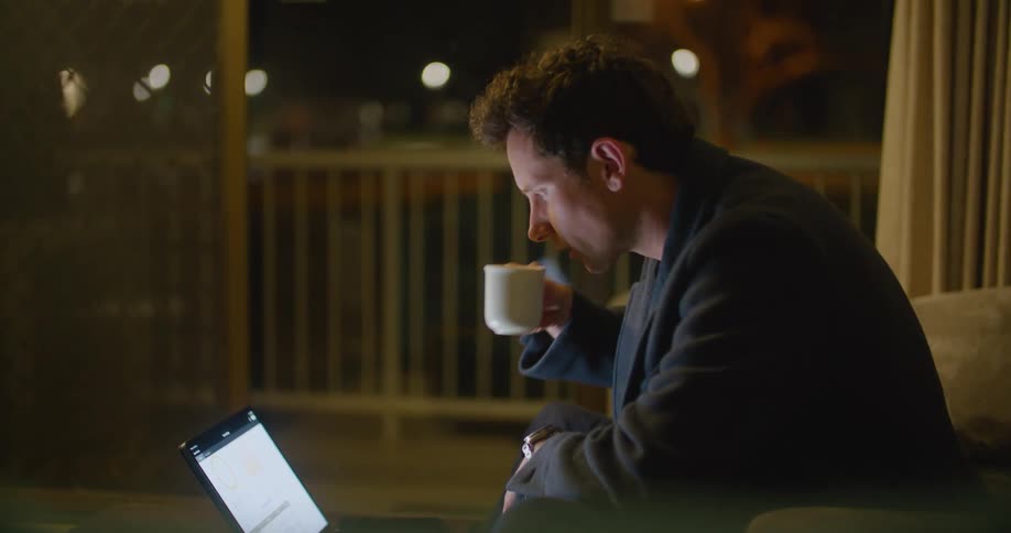 Man wearing thick robe sits on sofa drinking coffee and working on laptop