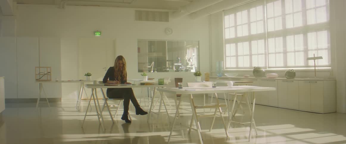 Blonde woman with a black blouse and green pants makes a drawing in a room