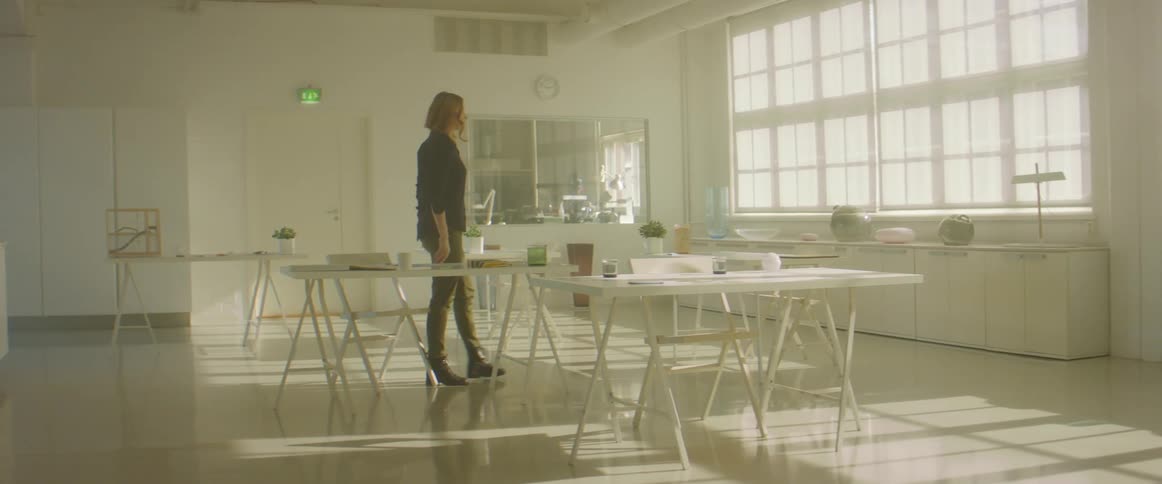 Blonde woman with a black blouse and green pants makes a drawing and stands up to look out the windows in a room
