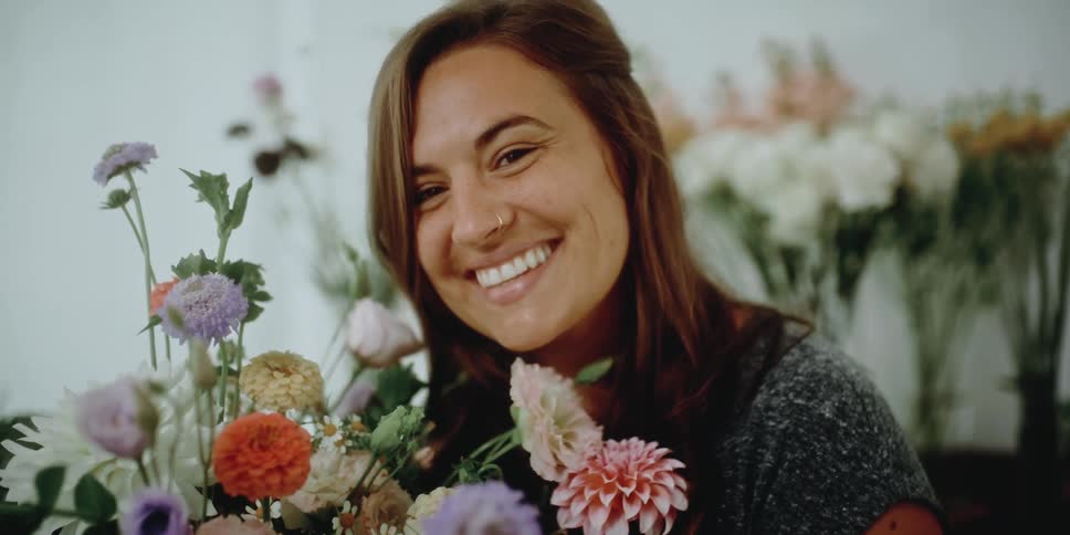 A woman smells a bouquet of flowers in a flower shop and lifts her head smiling and laughing