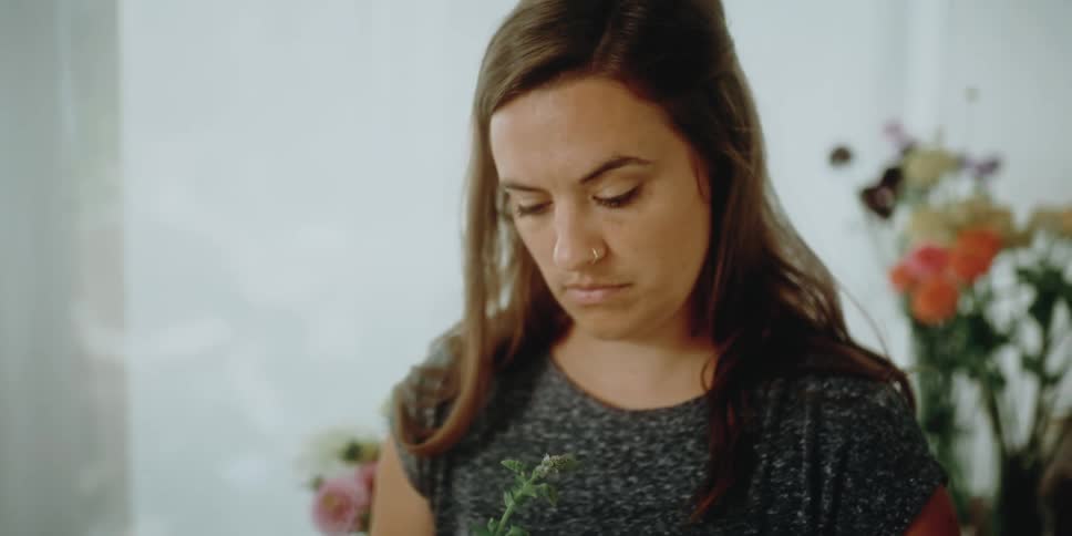 A woman in a room arranging flowers with a wall and plants in the background