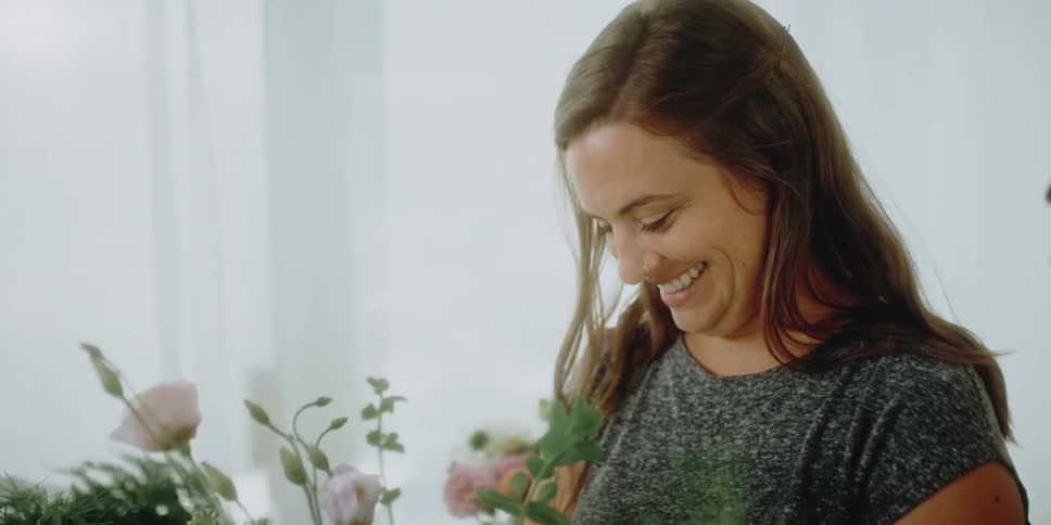 A woman in a room arranging flowers with a wall and plants in the background