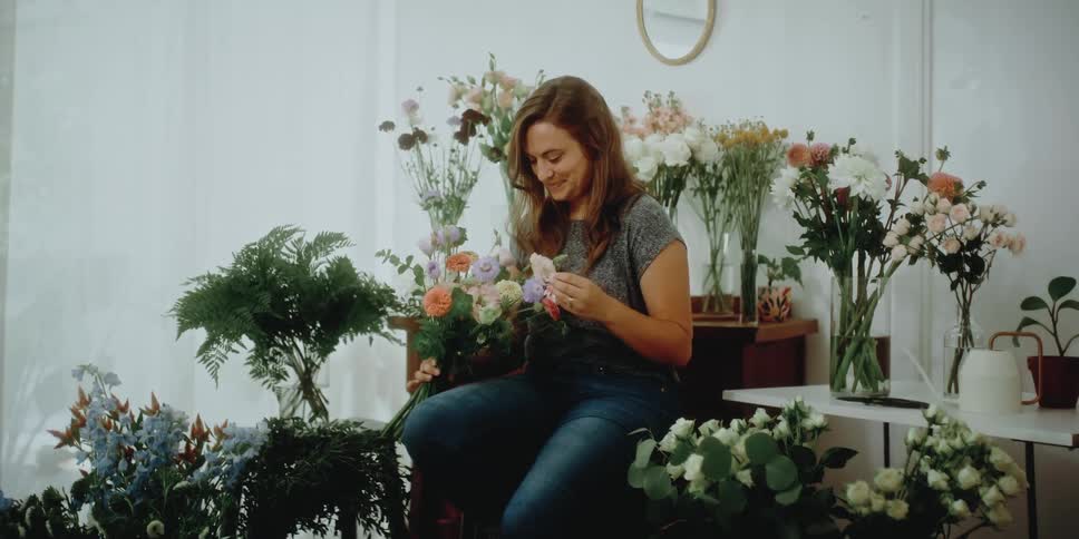 A smiling woman examines flowers in a bouquet while sitting indoors surrounded by house plants during the day