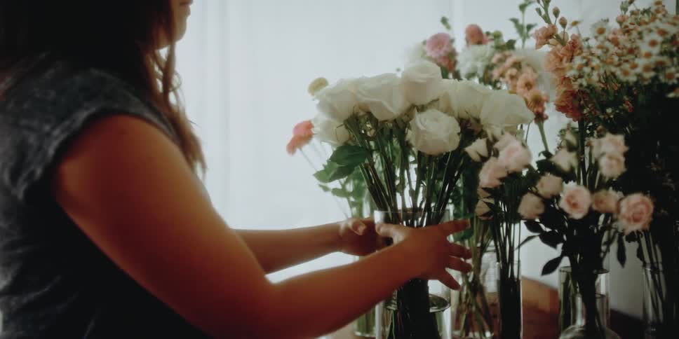 A florist moves a vase of white roses to a table covered with flower arrangements