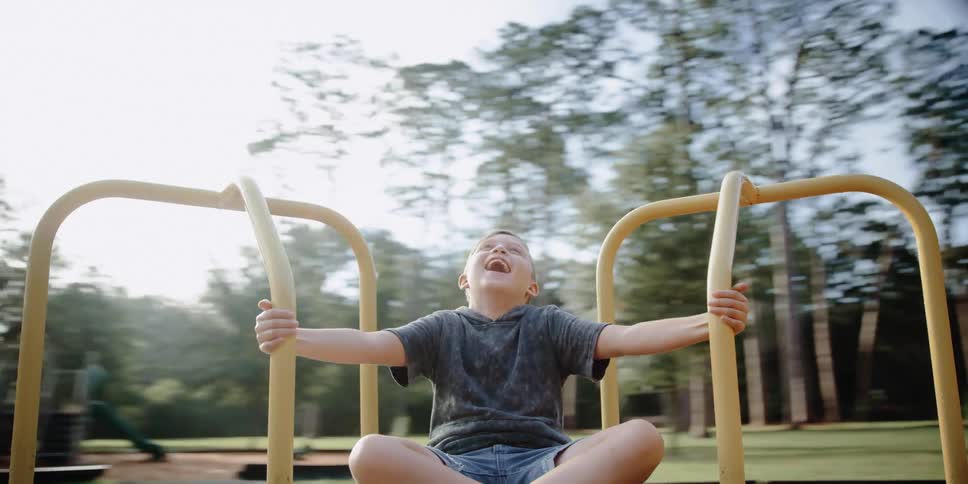 A boy playing on a merry go round with trees the sky and clouds in the background