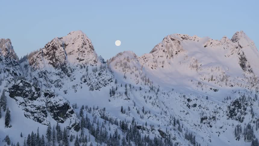 A bright and full moon traverses across a clear sky between the jagged peaks of snow covered mountains