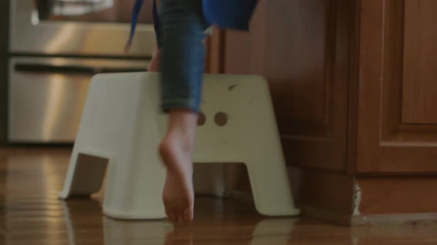 A child runs to a stool and steps on it to get something on a kitchen counter