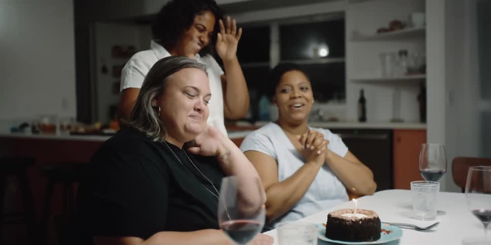 Three women sitting at a table with a cake with a candle and flame