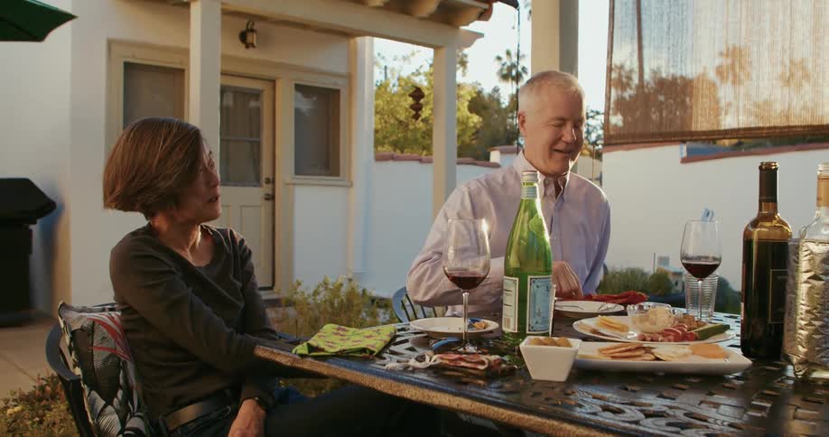 Couple sits in courtyard laughing and talking at table with food and wine