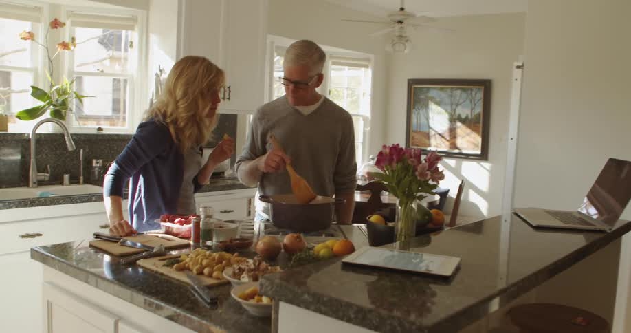 A man in a kitchen at home stirring food in a pot talks to his wife who is tasting an ingredient during the day