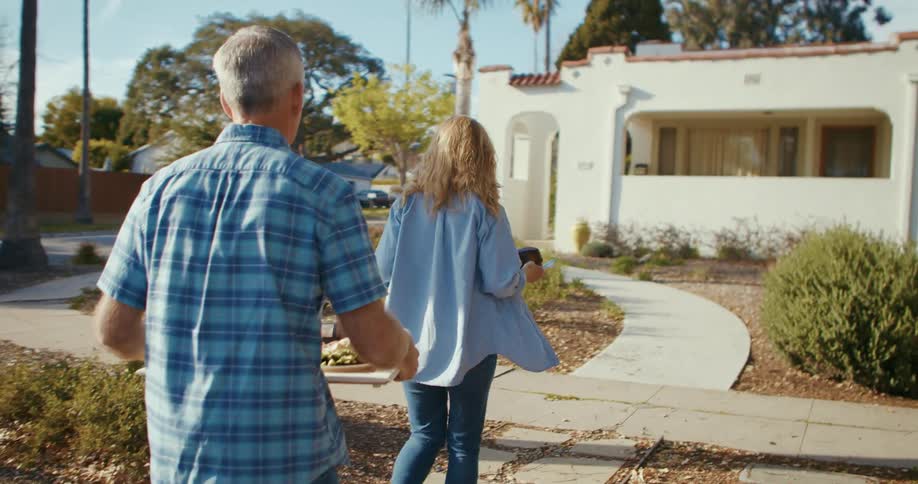 A man and a woman carrying trays of food walk towards a house in a neighborhood during the day