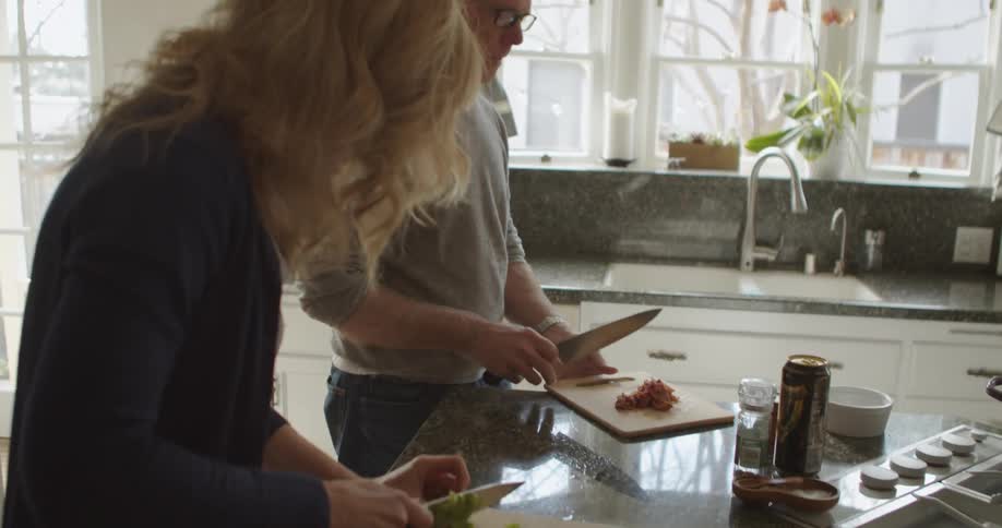 A man and a woman in the kitchen with the man adding meat into a pot on a stove