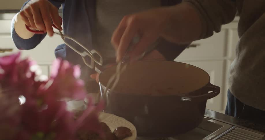 Two people in a kitchen at home using tongs to plate food from out of a pot during the day