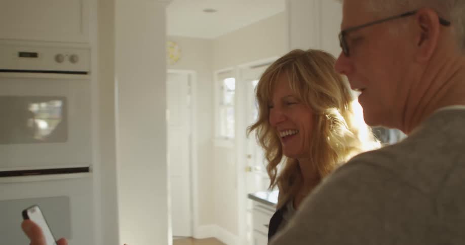 Man wearing glasses and grey sweatshirt and blonde woman laugh looking at smartphone while cooking in the kitchen