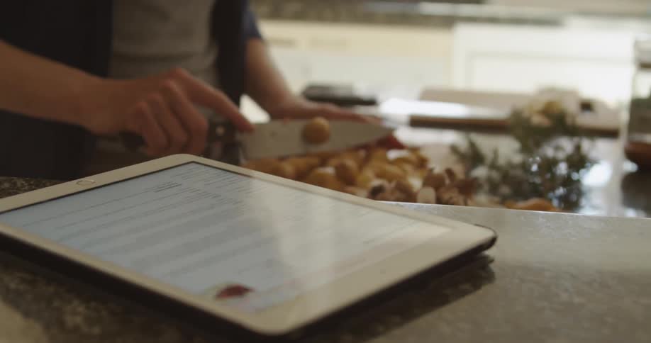 A woman prepares food in a kitchen with a tablet displaying a recipe on the counter next to her