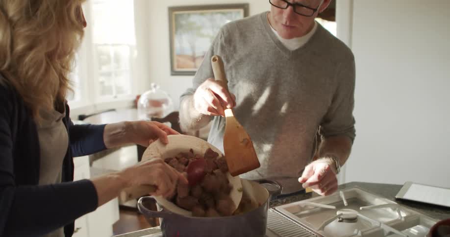 A man and woman pour roast beef from a bowl into a pot on an oven in a home kitchen during the day