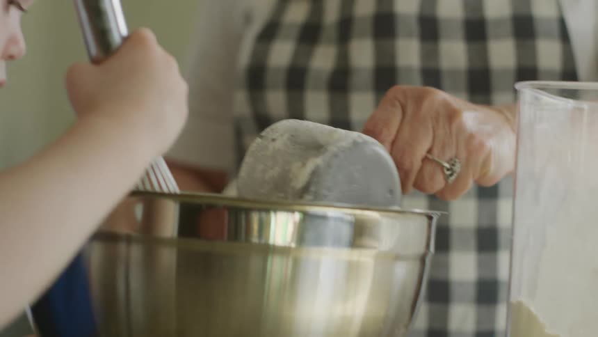 Female child stirs ingredients in metal mixing bowl as the grandmother uses a measuring cup to pour flour into the bowl