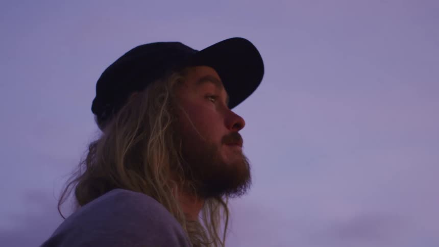 Side view of blonde man with beard long hair and black cap looks around at dusk